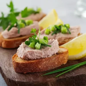 Toasts à la mousse de jambon disposés sur un plateau en bois avec herbes fraîches et citron
