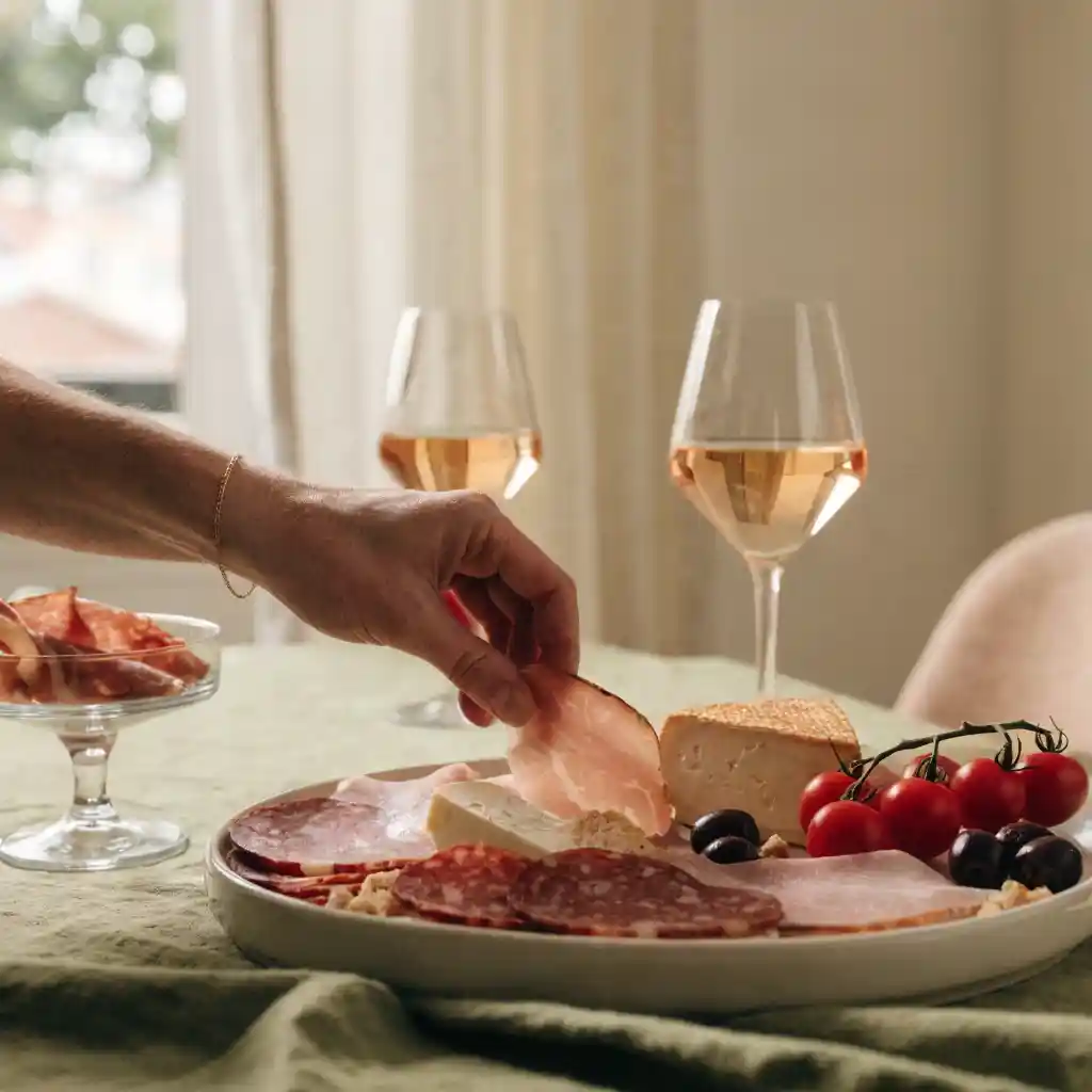 Planche de charcuteries et fromages servie à l'apéritif, une main prenant une tranche de jambon cru